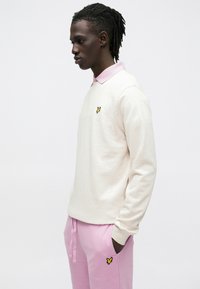 Világos krémszínű, hosszú ujjú polo rózsaszín gallérral és egy kis sárga pillangó logóval, rózsaszín szabadidőnadrággal, amelyen ugyanaz a logó található.