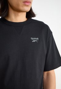 Melna kokvilnas t-krekls ar apaļu kakla izgriezumu un īsām piedurknēm. Uz kreisā krūts pusē ir neliels pelēks Reebok logotips.