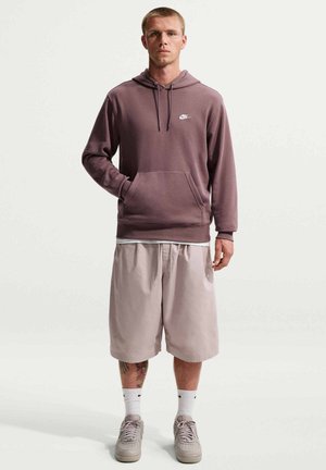 Młody mężczyzna stoi, ubrany w fioletowo-różową bluzę z kapturem Nike, jasnobeżowe krótkie spodenki do kolan, białe skarpetki Nike i beżowe sneakersy na jednolitym tle.