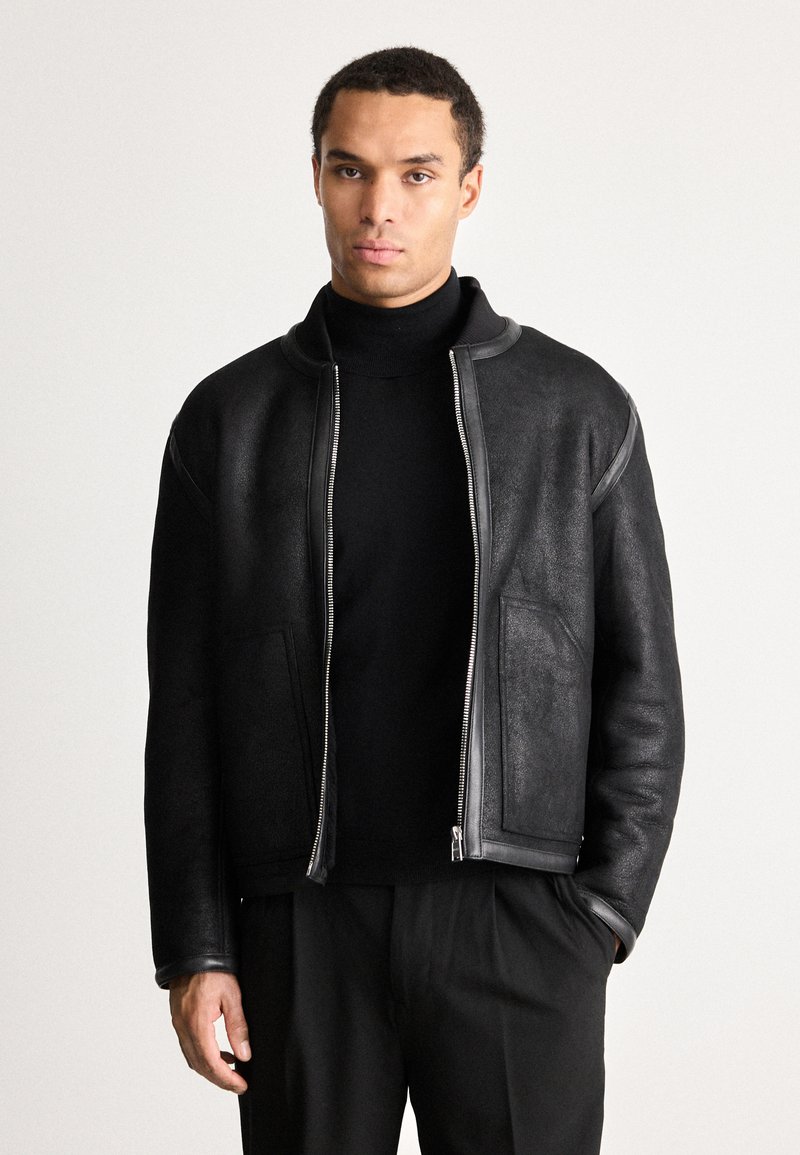 Hombre con chaqueta de cuero negra con cremallera sobre un suéter de cuello alto negro y pantalones negros, de pie contra un fondo claro y liso.