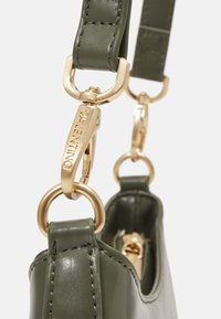 Bolso de cuero verde con accesorios dorados, incluyendo un clip y los lazos. Textura suave con costuras precisas y una forma estructurada.