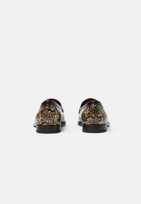 Zapatos de cuero negro con intrincados patrones abstractos dorados en la parte posterior. El interior tiene un forro burdeos que contrasta. Suela plana y sólida.
