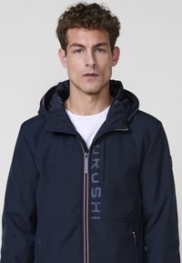 Chaqueta navy con cremallera y capucha, hecha de un tejido suave. Cuenta con un logo visible en el lado izquierdo y acentos de cremallera en contraste.