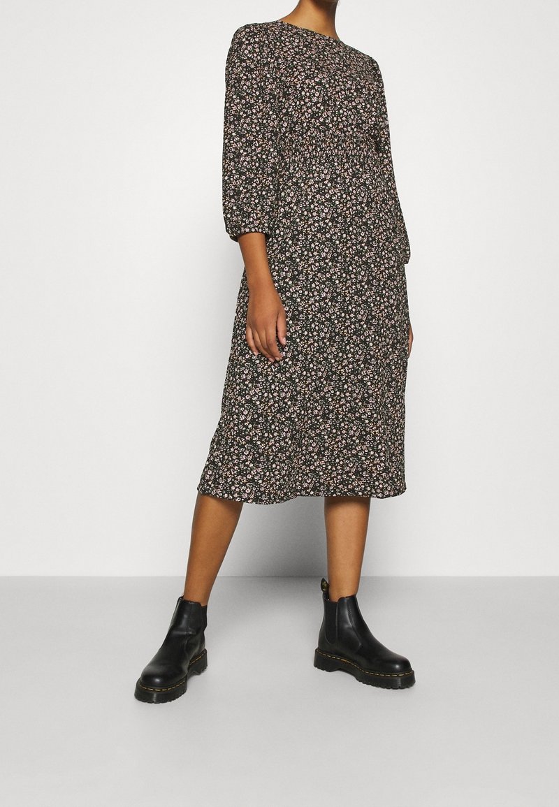Femme portant une robe midi noire à petit imprimé floral et manches bouffantes, associée à des bottines noires, debout sur un sol gris.