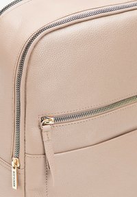 Béžová kožená taška s texturovaným povrchem, zlatými zipy a přední kapsou se zipem pro další úložný prostor.