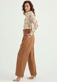 Weiße Bluse mit abstrakten orangefarbenen und braunen Mustern, kombiniert mit einer hoch taillierten braunen weiten Hose. Die Schuhe sind nude hohe Absätze.