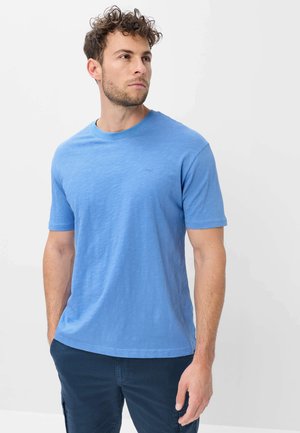 Mann mit kurzen lockigen Haaren, der ein hellblaues T-Shirt mit kurzen Ärmeln und dunkelblaue Hose trägt und nach links schaut.