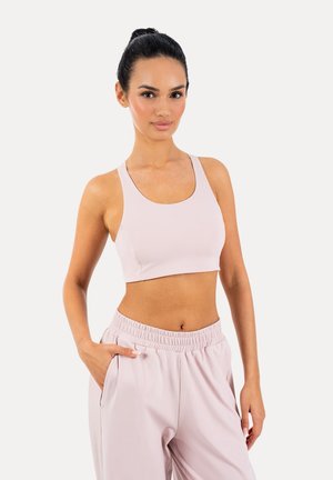 Femme portant un soutien-gorge de sport rose clair et un jogging taille haute assorti, debout avec une main dans la poche sur fond blanc.