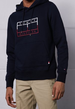 Mężczyzna w granatowej bluzie z kapturem Tommy Hilfiger z logo na piersi oraz beżowych spodniach na jednolitym szarym tle.