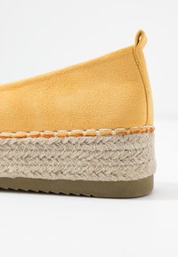 Gul ruskindsespadrille med en juteindpakket platform. Den har en tekstureret overdel og en solid gummisål, prydet med orange syninger.