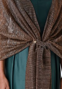 Shawl metallico marrone con un design tessuto e testurizzato, legato davanti con un bottone decorativo. Indossato sopra un abito verde scuro.