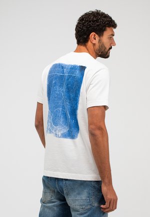 Mann trägt weißes T-Shirt mit abstraktem blauen Grafikdesign auf dem Rücken, kombiniert mit ausgewaschenen blauen Jeans, steht vor einfarbigem hellem Hintergrund.