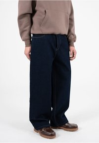 Jeans anchos de mezclilla azul oscuro con costuras visibles, combinados con una sudadera con capucha marrón y mocasines marrones con un detalle de cadena.