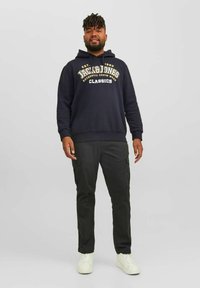 Marineblaue Kapuzenjacke mit Logo, kombiniert mit dunklen Cargo-Hosen mit Seitenentaschen. Stylisch mit weißen Sneakers, vor einem schlichten Hintergrund stehend.