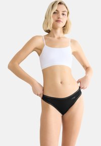 Mustat bikinihousut, joissa on pitsireunus ja logo, yhdistettynä valkoiseen, ohuilla olkaimilla varustettuun crop topiin. Sileä pinta ja istuva malli.