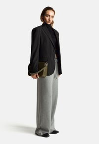 Czarna marynarka z dwoma guzikami, połączona z szarymi, szerokimi spodniami dresowymi. W dłoni trzymana jest fakturowana zielona torebka, a na nogach czarne buty.