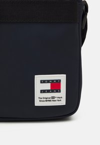 Borsa a tracolla navy, realizzata in materiale liscio, presenta una tracolla nera e un'etichetta rettangolare bianca con accenti rossi, bianchi e blu.