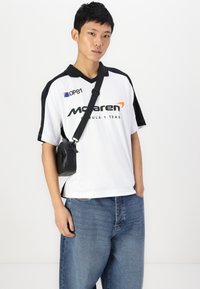 White short-sleeve shirt with black accents, featuring a collar and McLaren logo. Pair of loose blue jeans and a small black bag.