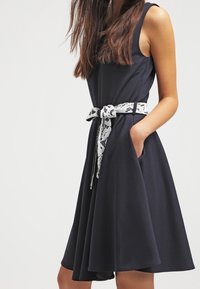 Abito senza maniche di colore blu navy con gonna a ruota e tasche laterali. La vita è accentuata da una cintura in pizzo bianco.