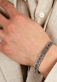 Bracciale in metallica argento con motivo intrecciato e chiusura piatta con logo. Indossato su una mano contro un tessuto di colore chiaro.