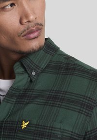 Camicia di flanella verde a quadri con strisce nere, dotata di colletto button-down e logo di un'aquila ricamato giallo sul petto.