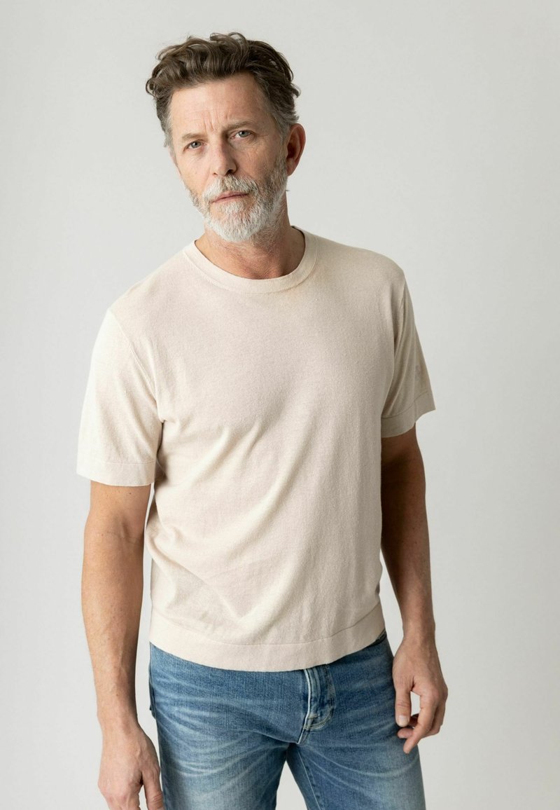 Homme à la barbe grise portant une chemise beige à manches courtes et un jean bleu, debout devant un fond clair uni.