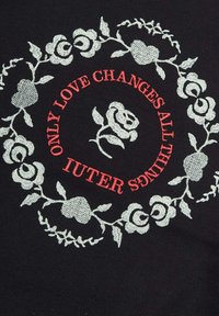 Tessuto nero con ricamo floreale bianco circolare e testo rosso che recita: "SOLAMENTE L'AMORE CAMBIA TUTTE LE COSE." Design centrale di una rosa.