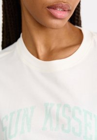 T-shirt en coton blanc avec l'inscription "SUN KISSED" en imprimé vert menthe délavé. Encolure ronde et texture lisse, avec des détails de couture visibles.