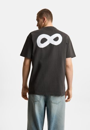 Mężczyzna w czarnym t-shircie z dużym białym symbolem nieskończoności na plecach, w połączeniu z jasnoniebieskimi dżinsami, stoi na tle jednolitego tła.