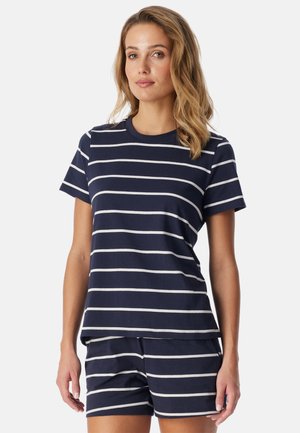 Vrouw draagt een marineblauw topje met korte mouwen en korte broek met witte horizontale strepen, staand tegen een effen achtergrond.