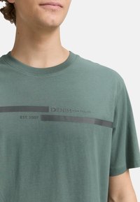 Grøn bomulds T-shirt med korte ærmer, der har et grafisk design i mørkegrå, inklusive brandnavn og etableringsdato.