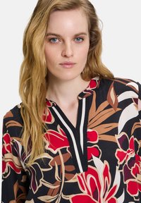 Bloemenblouse in zwart, met rode en beige patronen. Het ontwerp heeft een V-hals met gestreepte accenten en een zachte, gestructureerde stof.