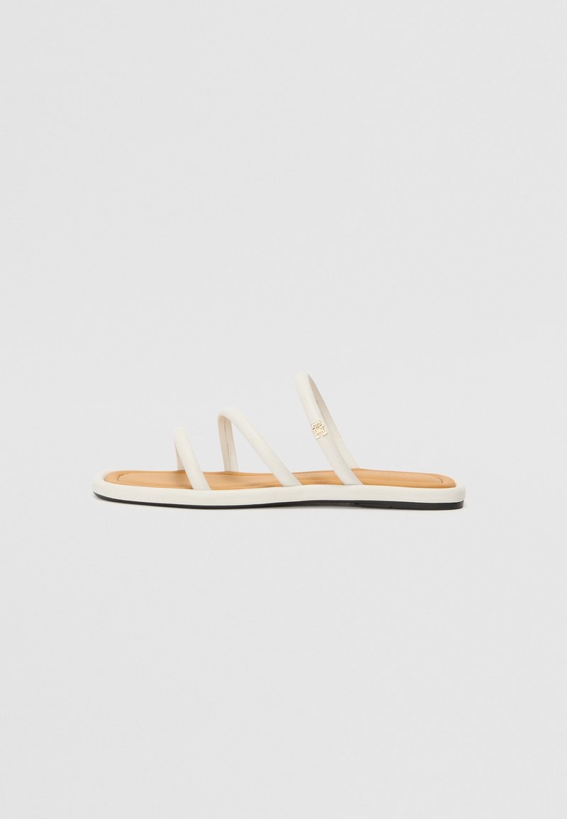 Sandale beige minimaliste avec trois fines lanières et une semelle plate, ornée d'un petit logo doré sur la lanière centrale.