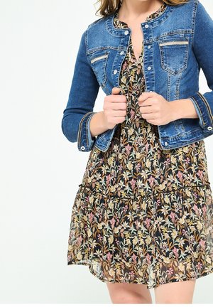 Vrouw draagt een blauwe spijkerjas over een zwarte bloemenjurk, houdt met beide handen de randen van de jas vast, tegen een effen lichte achtergrond.