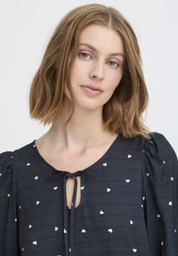 Blusa azul oscura con pequeños patrones de corazones blancos. Cuenta con un escote redondo, cierre con lazo y mangas abullonadas, hecha de tejido texturizado.