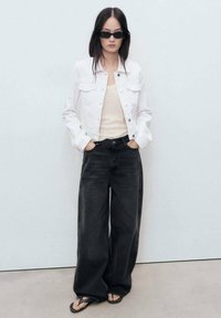 White denim jacket, beige top, black wide-leg jeans, and black sandals with studs. Model has long dark hair and sunglasses. Plain background.