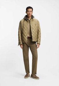 Uomo che indossa una giacca trapuntata color sabbia, polo beige, pantaloni marroni e scarpe marroni, in piedi davanti a uno sfondo bianco.