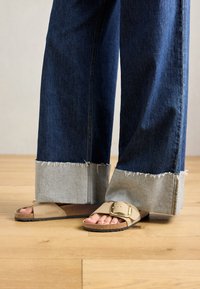 Noge v bež Birkenstock sandalih z zlatimi zaponkami, v kombinaciji s temno modrimi kavbojkami, zavihanimi pri robovih, stojijo na lesenem tleh.