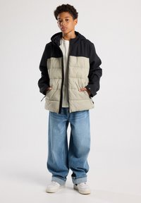 Padded jacket with a black upper and light grey lower portion, featuring a hood, zip closure, and two side pockets. Worn with loose blue jeans.