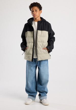 Chaqueta acolchada con parte superior negra y la inferior en gris claro, con capucha, cierre de cremallera y dos bolsillos laterales. Combinada con unos jeans azules holgados.