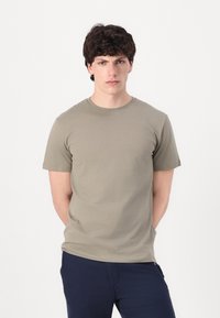 T-shirt κοντομάνικος, με στρογγυλή λαιμόκοψη, σε ανοιχτό ελαιόλαδο από βαμβακερό ύφασμα. Διαθέτει εφαρμοστό κόψιμο και λείο ύφασμα, φορεμένος με ναυτικές παντελόνες.