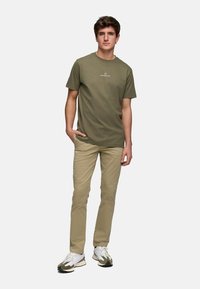 Camiseta verde oliva con logotipo, combinada con pantalones beige claros. El modelo está en una pose casual, vistiendo zapatillas de correr blancas y verdes.