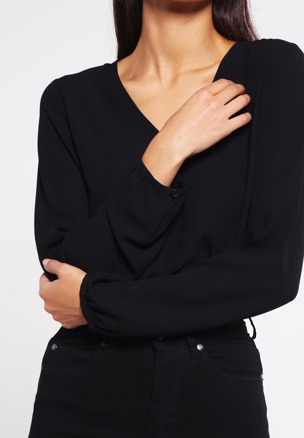 Femme portant un chemisier noir à manches longues avec col en V et poignets boutonnés, croisant les bras devant son torse.