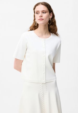 Kobieta o falowanych włosach nosi biały krótki top na guziki i dopasowaną spódnicę z ażurowymi detalami, dopełnioną dużymi złotymi kolczykami kołami.