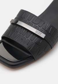 Sandale noire à glissière avec une bande texturée présentant des lettres en relief, un accent métallique DKNY et une zone des orteils noire brillante.