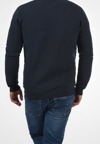 Tmavě modrý texturovaný svetr s žebrovanými manžetami a lemem, nošený přes světle modré džíny, zobrazuje zadní pohled a jemné horizontální vzory.