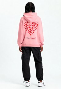 Felpa rosa con un motivo a cuore composto da cuori rossi sul retro, con la parola "SWEET" in corsivo nero. Abbinata a pantaloni cargo neri.
