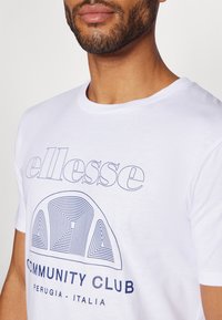 Balta medvilnė marškinėliai su navy spalvos grafiniu dizainu, kuriame yra tekstas „ellesse“ ir išlenktas raštas virš žodžių „COMMUNITY CLUB“ ir „PERUGIA - ITALIA“. 
