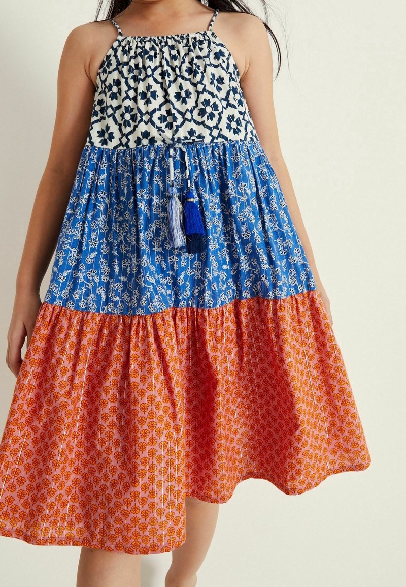 Robe sans manches à étages avec un haut floral blanc et marine, une partie centrale bleue avec des fleurs blanches, et un bas orange avec de petits motifs rouges.