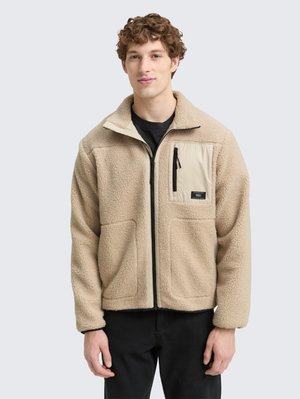 Junger Mann in beiger Fleecejacke mit Reißverschluss und Reißverschlusstasche auf der Brust sowie schwarzen Hosen vor einfarbigem Hintergrund.